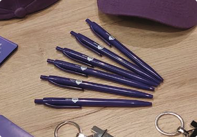 Set de bolígrafos promocionales morados personalizados con logo impreso por Bresspop, exhibidos junto a otros artículos de material POP como gorras y llaveros en una superficie de madera.